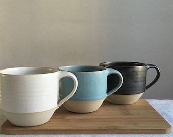Może przedstawiać: Trzy ceramiczne kubki w różnych kolorach: białym, jasnoniebieskim i ciemnoszarym. Kubki mają zaokrąglony kształt i uchwyt po prawej stronie. Są ułożone w rzędzie na drewnianej powierzchni.