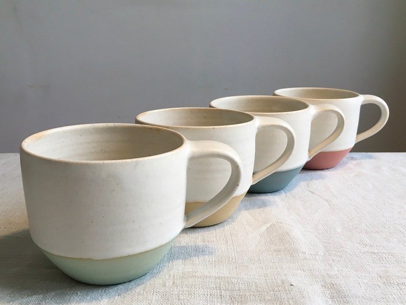May include: Four ceramic mugs with white interiors and colored rims. The mugs are lined up in a row, with the rim colors progressing from light blue to beige to pink.