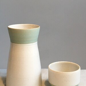 Może przedstawiać: Biały ceramiczny wazon z jasnozielonym paskiem wok&oacute;ł g&oacute;rnej części i mniejsza biała ceramiczna miska z jasnozielonym paskiem wok&oacute;ł dolnej części. Oba elementy znajdują się na białej powierzchni.