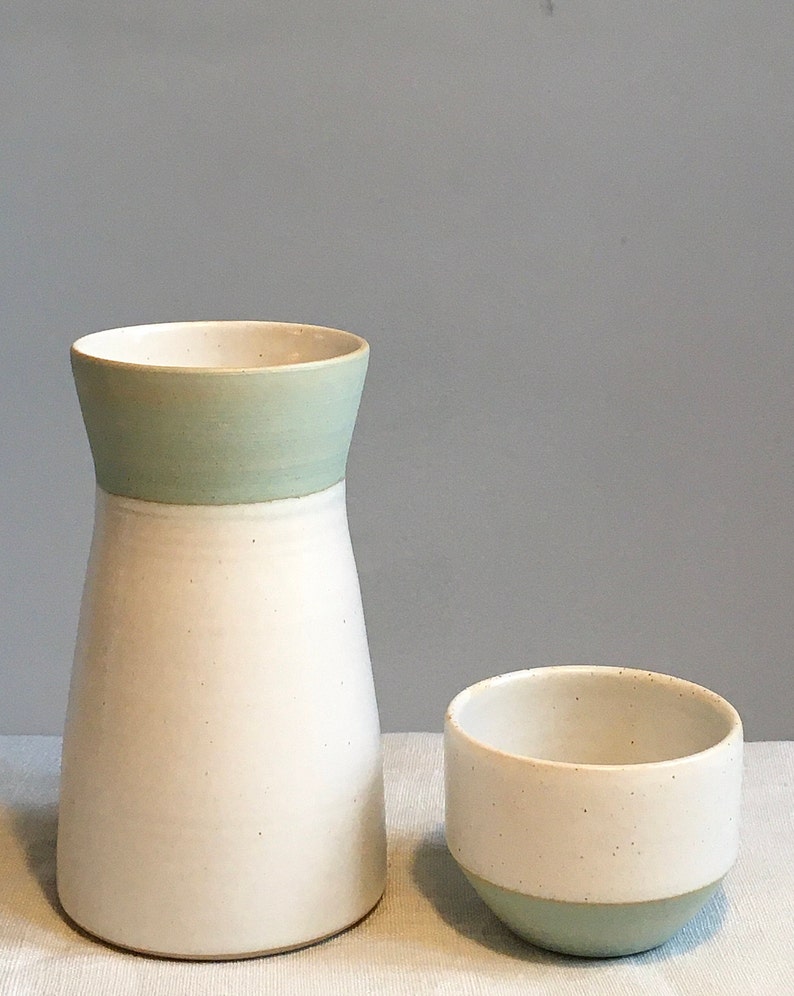 Może przedstawiać: Biały ceramiczny wazon z jasnozielonym paskiem wok&oacute;ł g&oacute;rnej części i mniejsza biała ceramiczna miska z jasnozielonym paskiem wok&oacute;ł dolnej części. Oba elementy znajdują się na białej powierzchni.