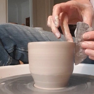 May include: A person is shaping a light brown ceramic mug on a pottery wheel using a tool. The mug has a rounded bottom and a wide opening.