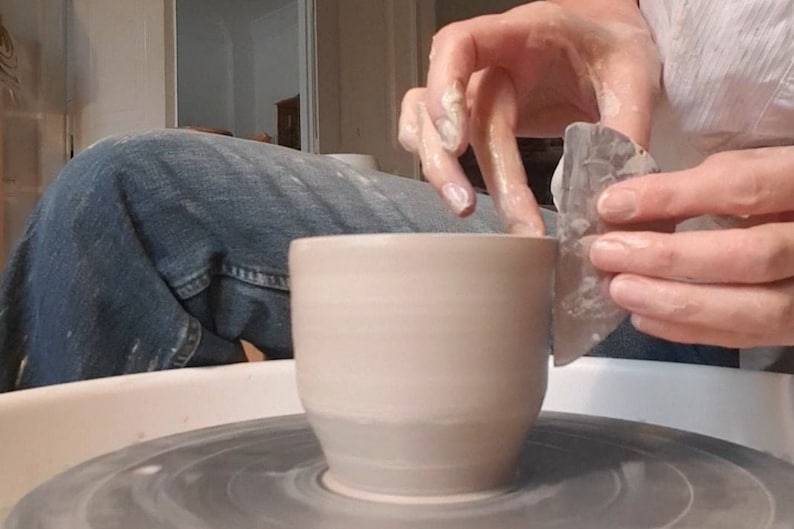 May include: A person is shaping a light brown ceramic mug on a pottery wheel using a tool. The mug has a rounded bottom and a wide opening.