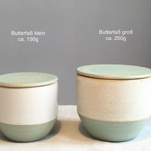 May include: Two ceramic butter dishes with lids. The smaller dish is labelled "Butterfa&szlig; klein ca. 150g" and the larger dish is labelled "Butterfa&szlig; gro&szlig; ca. 250g". Both dishes are white with a light green rim and lid.