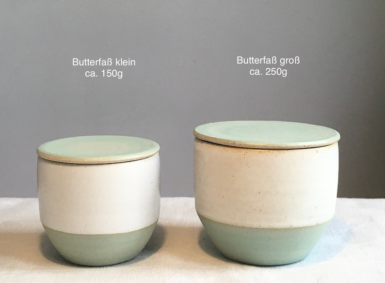 May include: Two ceramic butter dishes with lids. The smaller dish is labelled "Butterfa&szlig; klein ca. 150g" and the larger dish is labelled "Butterfa&szlig; gro&szlig; ca. 250g". Both dishes are white with a light green rim and lid.