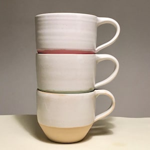 May include: Three white ceramic mugs stacked on top of each other. The top mug has a pink rim, the middle mug has a light green rim, and the bottom mug has a light brown rim.