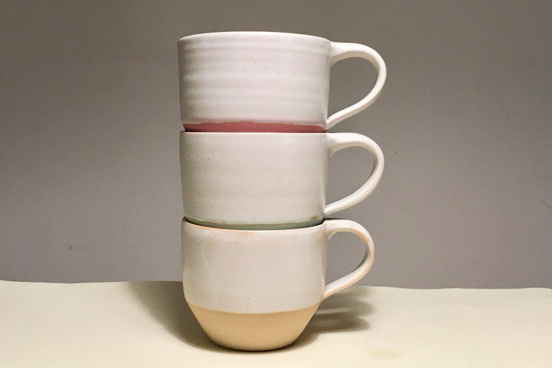 May include: Three white ceramic mugs stacked on top of each other. The top mug has a pink rim, the middle mug has a light green rim, and the bottom mug has a light brown rim.