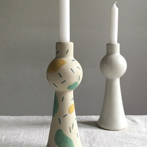 Op de afbeelding: Twee keramische kaarsenstandaards, een met een witte glazuur en de andere met een witte glazuur en abstracte blauwe, groene en gele geschilderde ontwerpen. Beide standaards hebben een ronde bovenkant en een uitlopende basis.