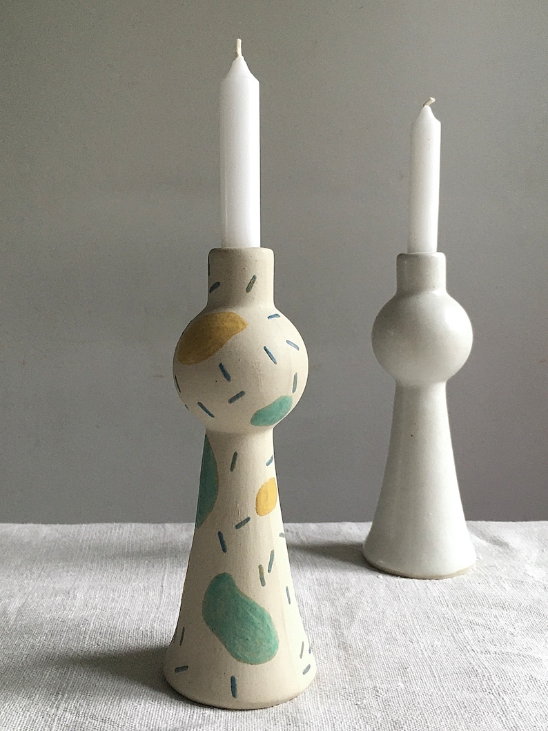 Op de afbeelding: Twee keramische kaarsenstandaards, een met een witte glazuur en de andere met een witte glazuur en abstracte blauwe, groene en gele geschilderde ontwerpen. Beide standaards hebben een ronde bovenkant en een uitlopende basis.