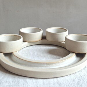 May include: A handmade, off-white ceramic serving dish with four small, round bowls arranged around a central circular tray. The pottery has a matte finish and a rustic, minimalist aesthetic, suitable for serving snacks or dips.