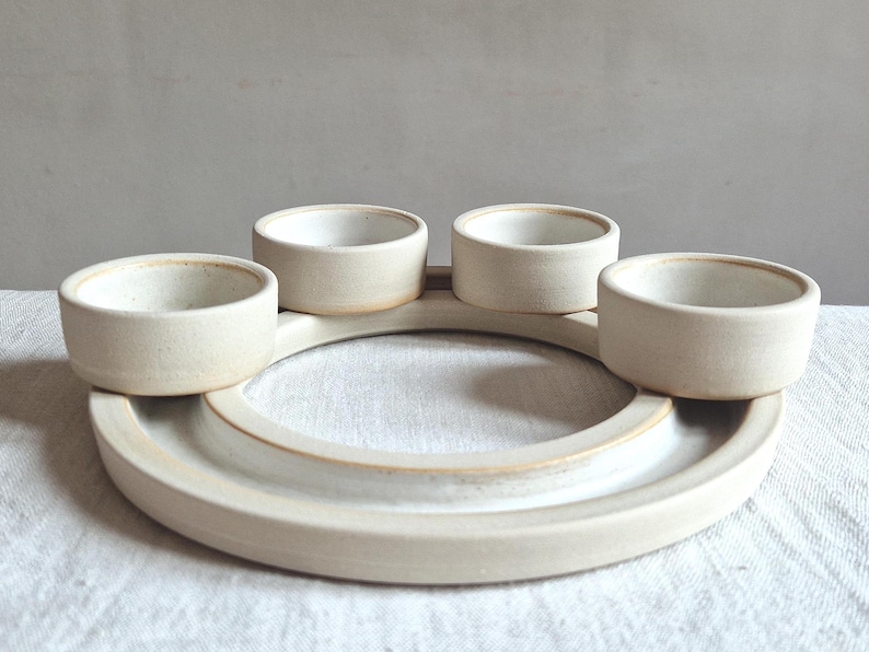 May include: A handmade, off-white ceramic serving dish with four small, round bowls arranged around a central circular tray. The pottery has a matte finish and a rustic, minimalist aesthetic, suitable for serving snacks or dips.