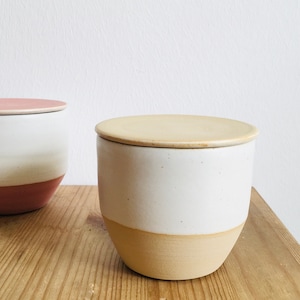 May include: Two ceramic storage containers with lids. The container on the left has a pink lid and a white body with a pink base. The container on the right has a beige lid and a white body with a beige base.