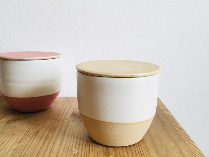 May include: Two ceramic storage containers with lids. The container on the left has a pink lid and a white body with a pink base. The container on the right has a beige lid and a white body with a beige base.