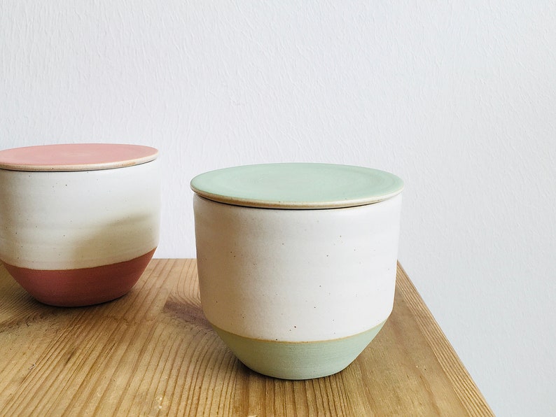 May include: Two ceramic storage jars with lids. The jar on the left has a pink lid and a white body with a pink band. The jar on the right has a light green lid and a white body with a light green band.