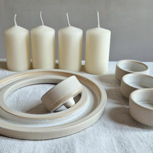 May include: A collection of cream-colored pillar candles, ceramic dishes, and small bowls. The candles are cylindrical with a wick at the top. The dishes and bowls are a light beige color, with a simple, minimalist design.