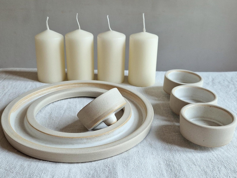 May include: A collection of cream-colored pillar candles, ceramic dishes, and small bowls. The candles are cylindrical with a wick at the top. The dishes and bowls are a light beige color, with a simple, minimalist design.