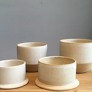 May include: Four white ceramic bowls with a light brown glaze on the bottom. The bowls are arranged in a pyramid shape on a wooden surface.