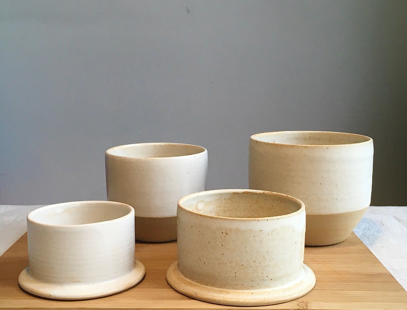May include: Four white ceramic bowls with a light brown glaze on the bottom. The bowls are arranged in a pyramid shape on a wooden surface.