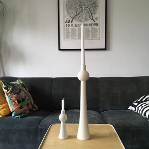 Op de afbeelding: Twee witte keramische kaarsenstandaards in de vorm van de Berliner Fernsehturm. De grotere standaard is hoger dan de kleinere. De standaards staan op een houten tafel voor een grijze bank.