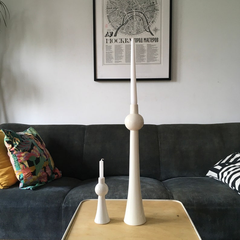 Op de afbeelding: Twee witte keramische kaarsenstandaards in de vorm van de Berliner Fernsehturm. De grotere standaard is hoger dan de kleinere. De standaards staan op een houten tafel voor een grijze bank.