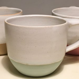 May include: Three ceramic mugs with different colored bands around the bottom. The mugs are white with a speckled glaze. The bands are light brown, light green, and light pink.