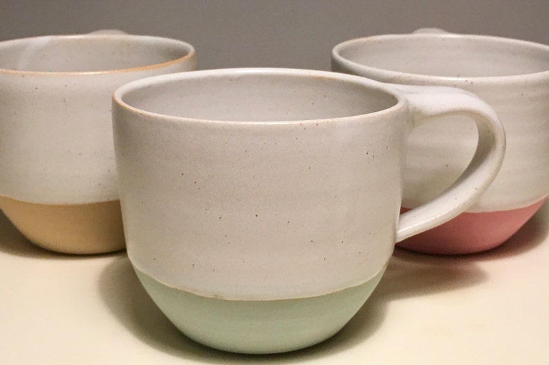 May include: Three ceramic mugs with different colored bands around the bottom. The mugs are white with a speckled glaze. The bands are light brown, light green, and light pink.