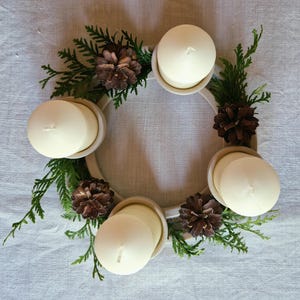 May include: A circular Advent wreath with four white candles, green sprigs, and brown pinecones. The candles are set in a cream-colored ring, creating a festive holiday centerpiece. The wreath is on a light-colored surface.