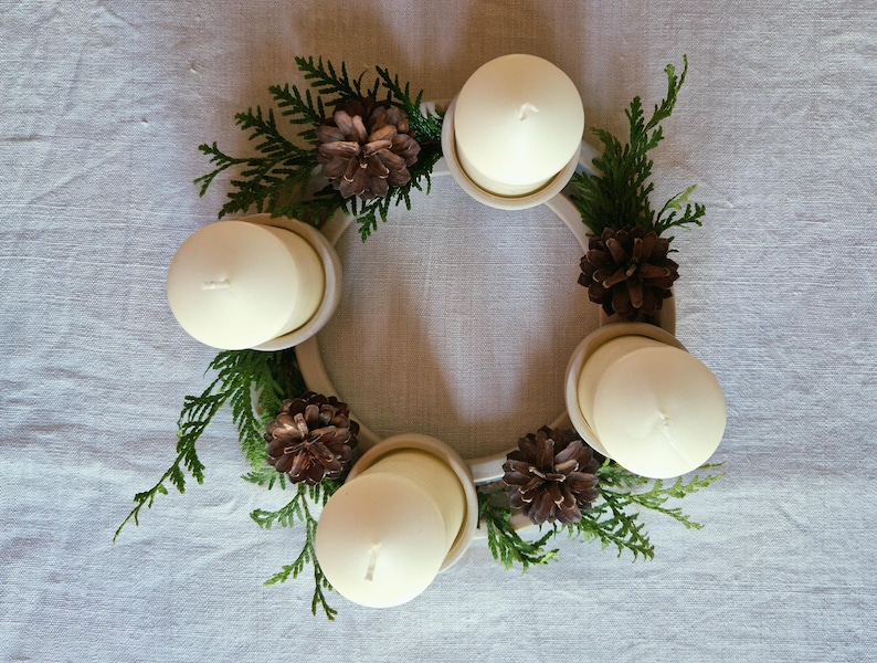 May include: A circular Advent wreath with four white candles, green sprigs, and brown pinecones. The candles are set in a cream-colored ring, creating a festive holiday centerpiece. The wreath is on a light-colored surface.