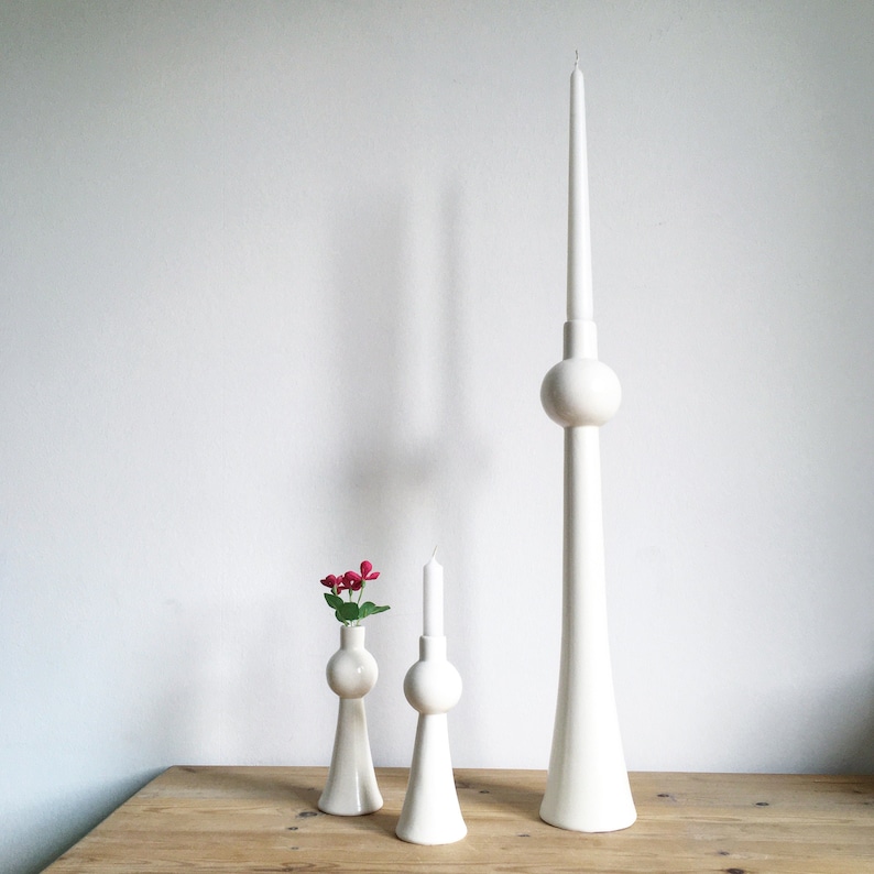 Op de afbeelding: Drie witte keramische kaarsenhouders in de vorm van een toren. De hoogste houder heeft &eacute;&eacute;n kaars, terwijl de twee kleinere houders elk &eacute;&eacute;n kaars hebben. De houders staan op een houten tafel met een klein boeket roze bloemen in de kleinste houder.