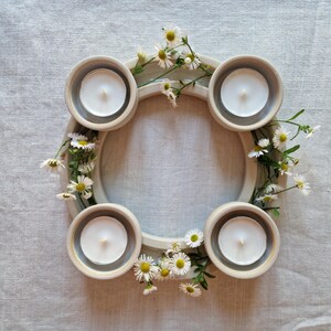 May include: A circular ceramic candle holder with four recessed areas, each holding a white tea light candle. The holder is decorated with small white and yellow flowers and green stems, creating a natural, decorative centerpiece.