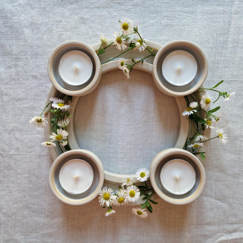 May include: A circular ceramic candle holder with four recessed areas, each holding a white tea light candle. The holder is decorated with small white and yellow flowers and green stems, creating a natural, decorative centerpiece.
