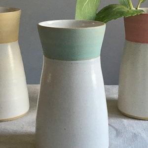 Może przedstawiać: Biały ceramiczny wazon z jasnoniebieskim pasem wok&oacute;ł g&oacute;rnej części. Wazon stoi na białym obrusie lnianym.