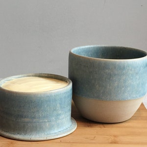 May include: A ceramic butter crock featuring a two-piece design with a light blue mottled glaze on the exterior. The base holds a block of cream-colored butter, while the taller, matching blue cup sits beside it, showcasing a smooth, unglazed base.