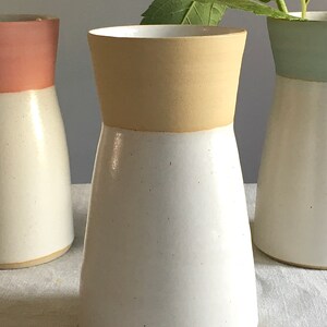 Może przedstawiać: Biały ceramiczny wazon z beżową g&oacute;rną krawędzią. Wazon ma szeroką podstawę i wąską szyjkę.