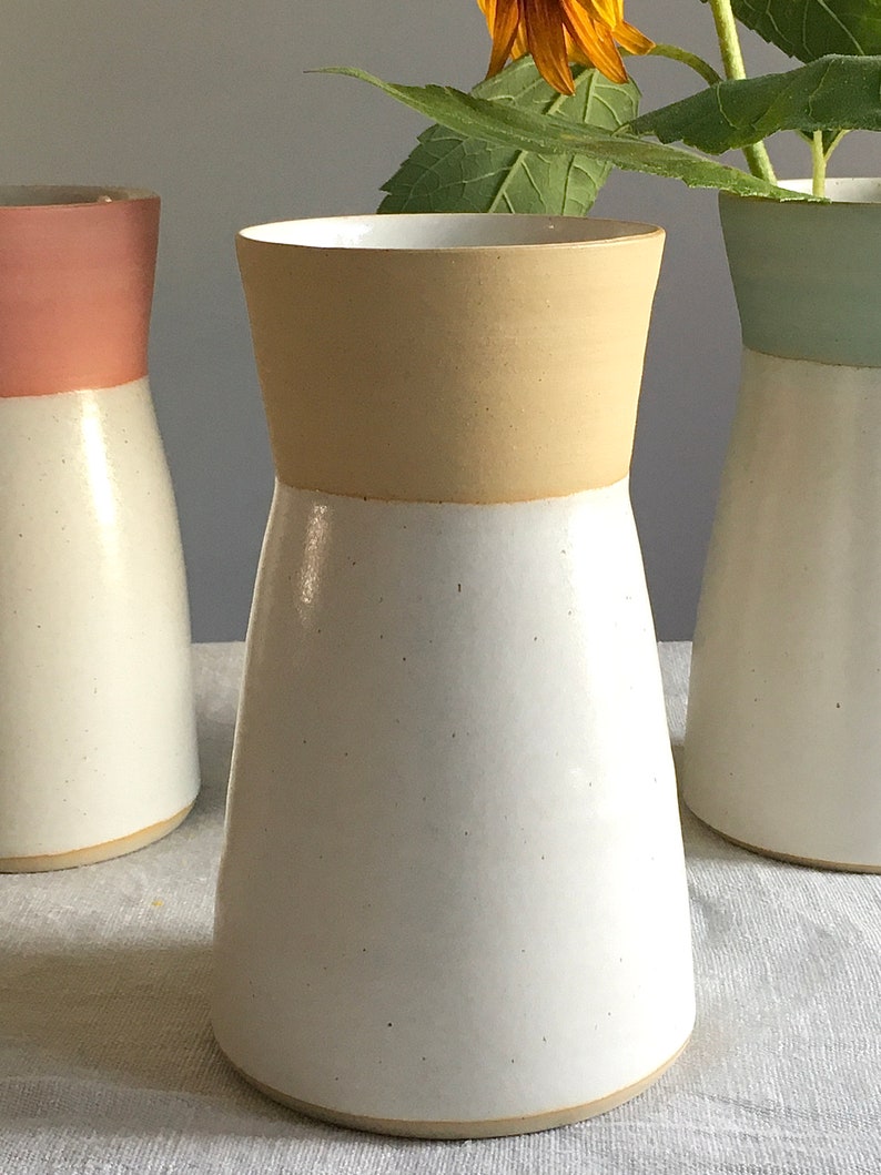 Może przedstawiać: Biały ceramiczny wazon z beżową g&oacute;rną krawędzią. Wazon ma szeroką podstawę i wąską szyjkę.