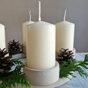 May include: A decorative arrangement featuring four ivory-colored pillar candles in ceramic holders. The candles are surrounded by pine cones and green sprigs, creating a natural, seasonal display. The arrangement is set on a light-colored, oval-shaped base.