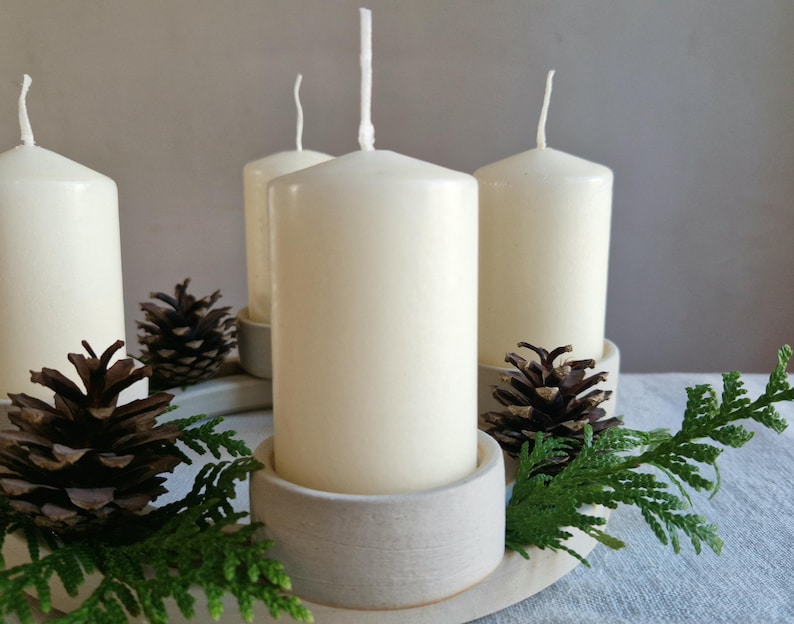 May include: A decorative arrangement featuring four ivory-colored pillar candles in ceramic holders. The candles are surrounded by pine cones and green sprigs, creating a natural, seasonal display. The arrangement is set on a light-colored, oval-shaped base.