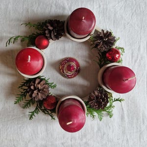May include: A festive Advent wreath with four red candles in white holders, surrounded by pinecones, red ornaments, and evergreen sprigs. The wreath is arranged on a white surface, creating a seasonal centerpiece.