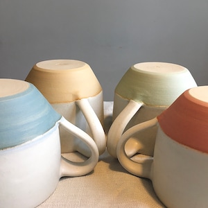 May include: Four ceramic mugs with white handles. Each mug is two-toned with a white base and a colored top. The colors are light blue, yellow, green, and pink.