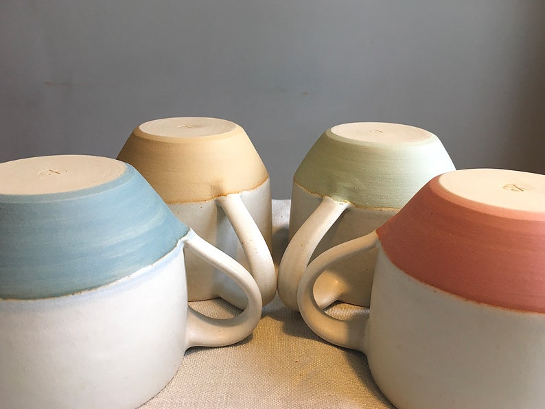 May include: Four ceramic mugs with white handles. Each mug is two-toned with a white base and a colored top. The colors are light blue, yellow, green, and pink.