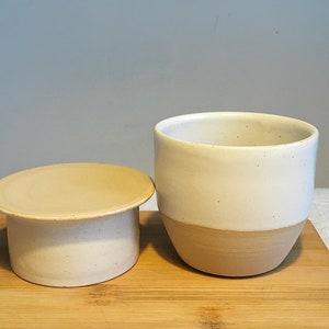 May include: A white ceramic butter dish with a lid. The dish has a speckled glaze and a beige base. The lid is a separate piece and has a smooth, beige top.