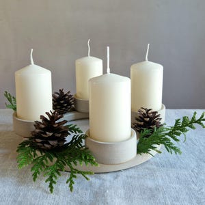 May include: A cream-colored Advent wreath with four white pillar candles. The candles are set in light gray holders and surrounded by pine cones and green sprigs. The wreath sits on a white surface.
