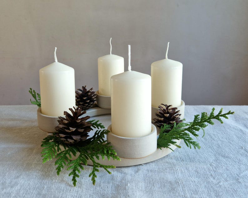 May include: A cream-colored Advent wreath with four white pillar candles. The candles are set in light gray holders and surrounded by pine cones and green sprigs. The wreath sits on a white surface.