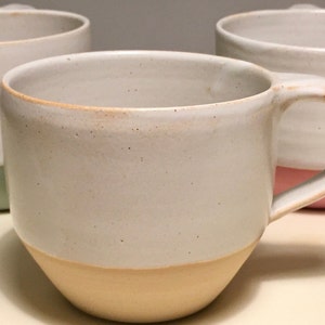 May include: Three ceramic mugs with a white glaze and a colored band around the bottom. The mugs are in a row, with the first mug having a light green band, the second mug having a light yellow band, and the third mug having a light pink band.