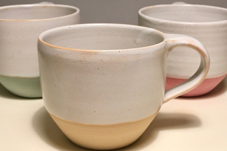May include: Three ceramic mugs with a white glaze and a colored band around the bottom. The mugs are in a row, with the first mug having a light green band, the second mug having a light yellow band, and the third mug having a light pink band.