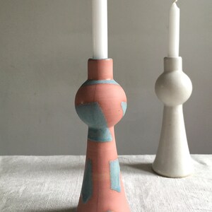 Op de afbeelding: Twee keramische kaarsenstandaards, een in wit en een met een geometrisch patroon in roze en blauw. Beide standaards hebben een ronde, bolle bovenkant en een uitlopende basis.