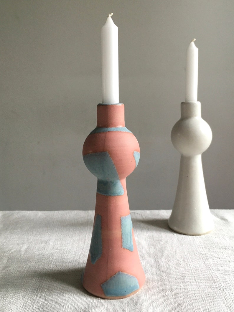 Op de afbeelding: Twee keramische kaarsenstandaards, een in wit en een met een geometrisch patroon in roze en blauw. Beide standaards hebben een ronde, bolle bovenkant en een uitlopende basis.