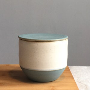 May include: A ceramic jar with a lid, featuring a white body and a light blue lid and base. The jar is sitting on a wooden surface.