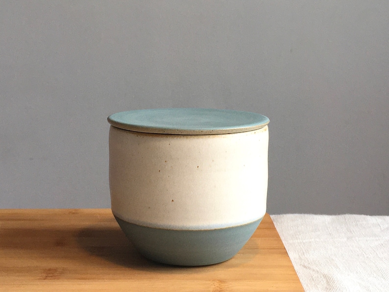 May include: A ceramic jar with a lid, featuring a white body and a light blue lid and base. The jar is sitting on a wooden surface.