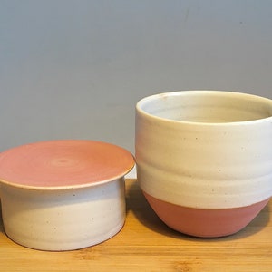 May include: A white ceramic butter dish with a pink lid. The dish has a white base and a pink rim. The lid has a pink top and a white base.