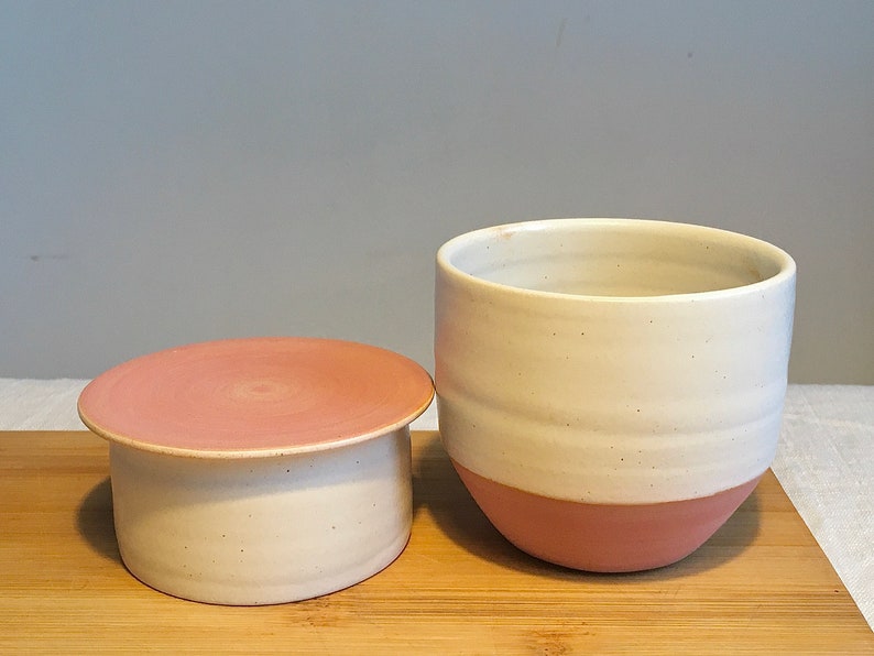 May include: A white ceramic butter dish with a pink lid. The dish has a white base and a pink rim. The lid has a pink top and a white base.